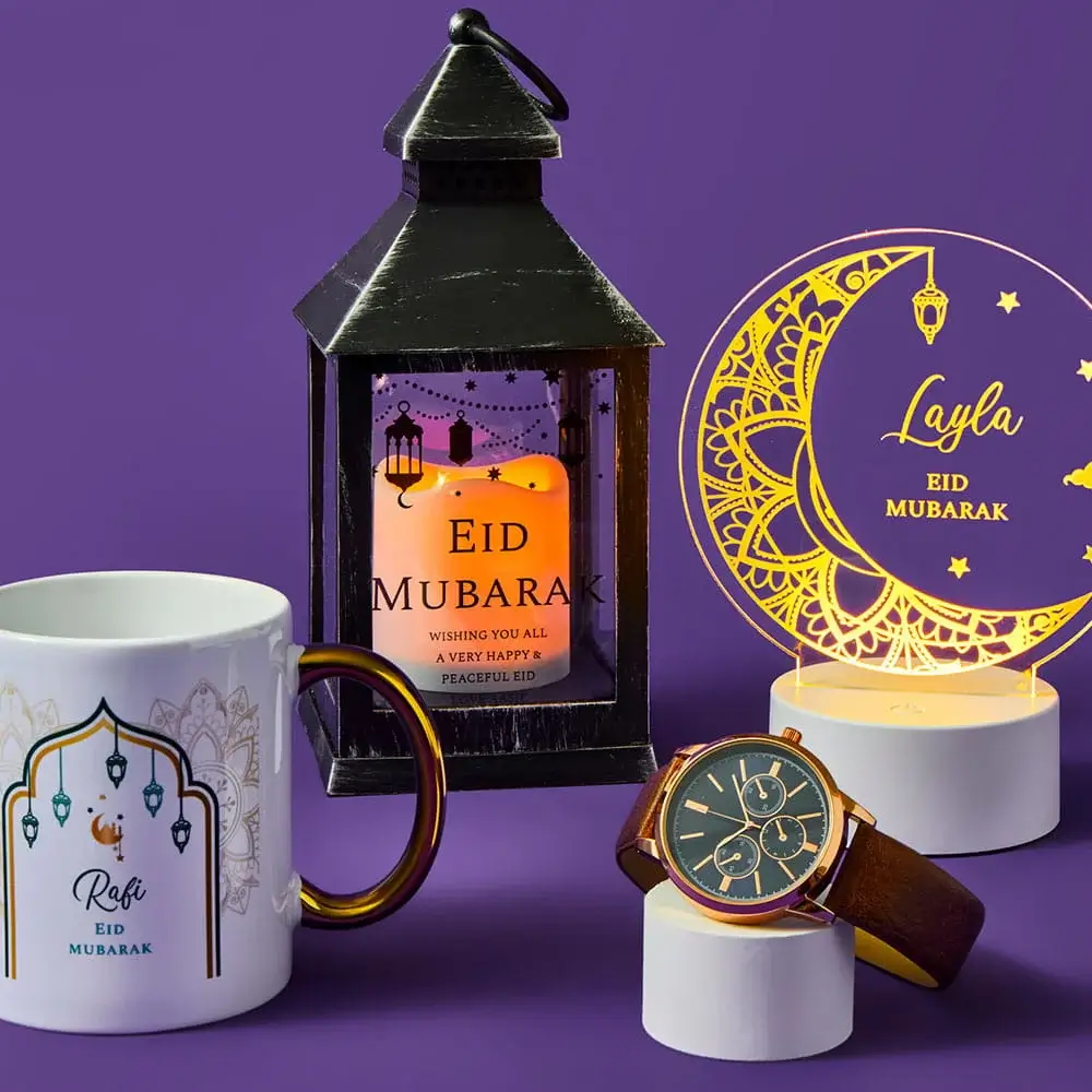 White Eid Mubarak mug, a glowing lantern with a candle, a personalised moon‑and‑stars light, and a brown‑strap wristwatch displayed against a purple background.